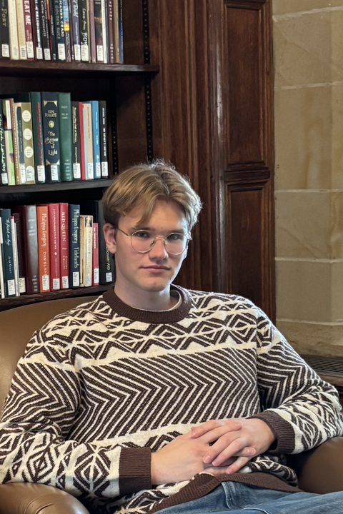 Jannik sitting in front of a bookcase wearing glasses and a patterned sweater.